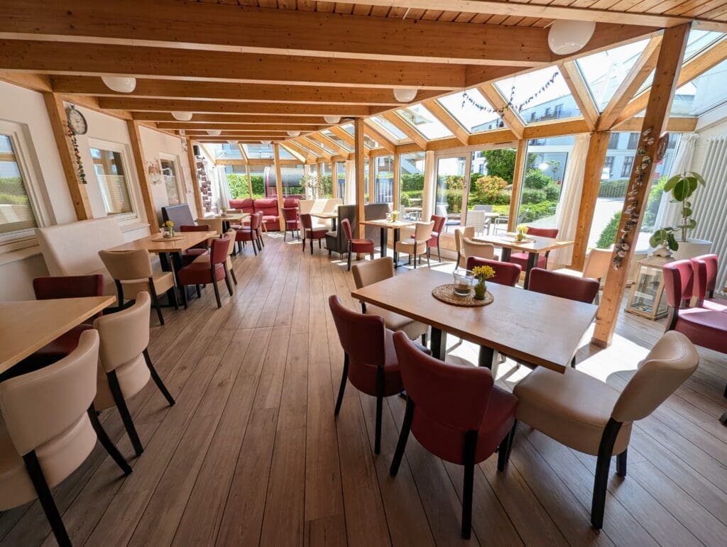 Helle Cafeteria mit Holzdecke und großen Fenstern, möbliert mit roten und beigen Polsterstühlen